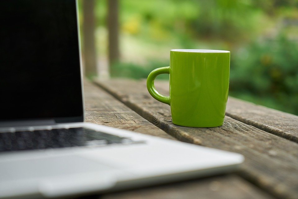 Ein Laptop draußen in der Natur auf einem verwitterten Holztisch. Daneben eine hellgrüne Kaffeetasse.